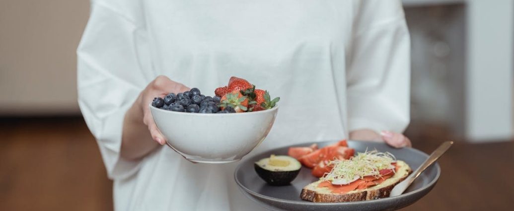 Person holding a bowl of fresh berries and a plate with avocado toast, displaying a healthy breakfast spread.